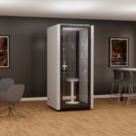 A modern office with a single-person phone booth featuring a frosted door and stool inside. The room has wooden flooring, gray walls with landscape photos, a small round table with high chairs, and a gray armchair.
