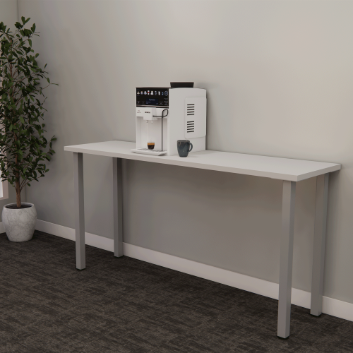 A modern coffee machine on a long white table dispenses a cup of coffee. A small plant in a white pot is placed on the floor next to the table. The room has a plain light gray wall and a dark carpeted floor.