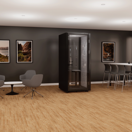 Modern office space with a black phone booth, gray walls, wooden flooring, and framed nature photos. There are gray lounge chairs with a black table on the left and a tall white table with stools on the right.