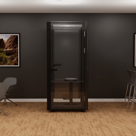 A modern, glass-enclosed phone booth with a stool inside stands in an office space with wooden flooring. The surrounding area features charcoal walls adorned with framed landscape photographs, and there are various chairs nearby.