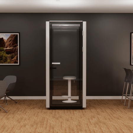 A small soundproof booth with a stool inside is placed against a dark wall in an office setting with wood flooring. Framed art hangs on the walls, and there are chairs nearby, including some bar stools.